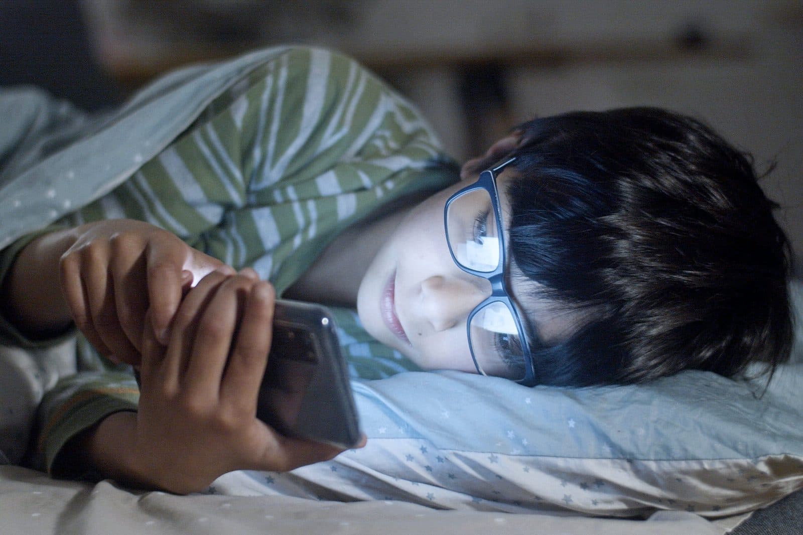 Stock photograph of a child lying in bed in the dark, lit by the screen of a phone held close to their face