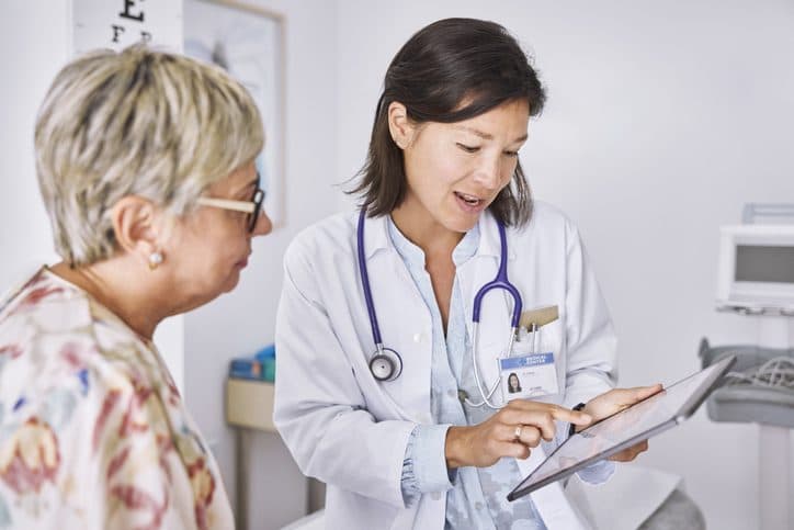 A clinician in a white coat holding a tablet in consultation with an older patient in a examination room