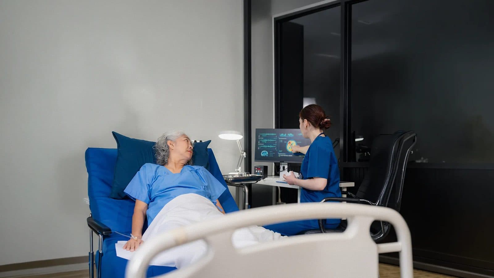 A patient in a hospital gown lying in a raised hospital bed while a clinician in scrubs works at a computer screen beside them.