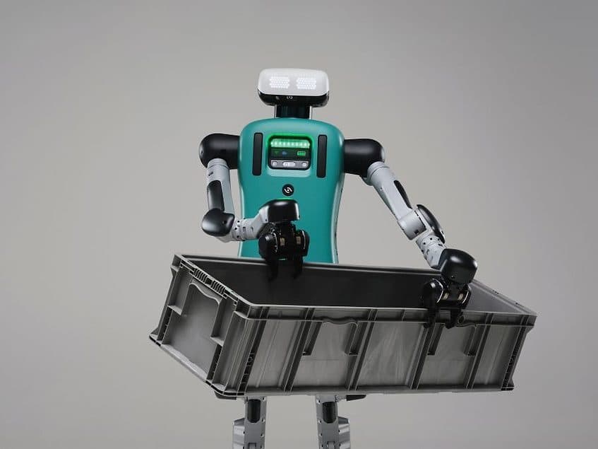 Agility Robotics 的 Digit 人形机器人,青绿色躯干和 LED 面部条,手持灰色塑料仓储周转箱,置于纯色工作室背景前
