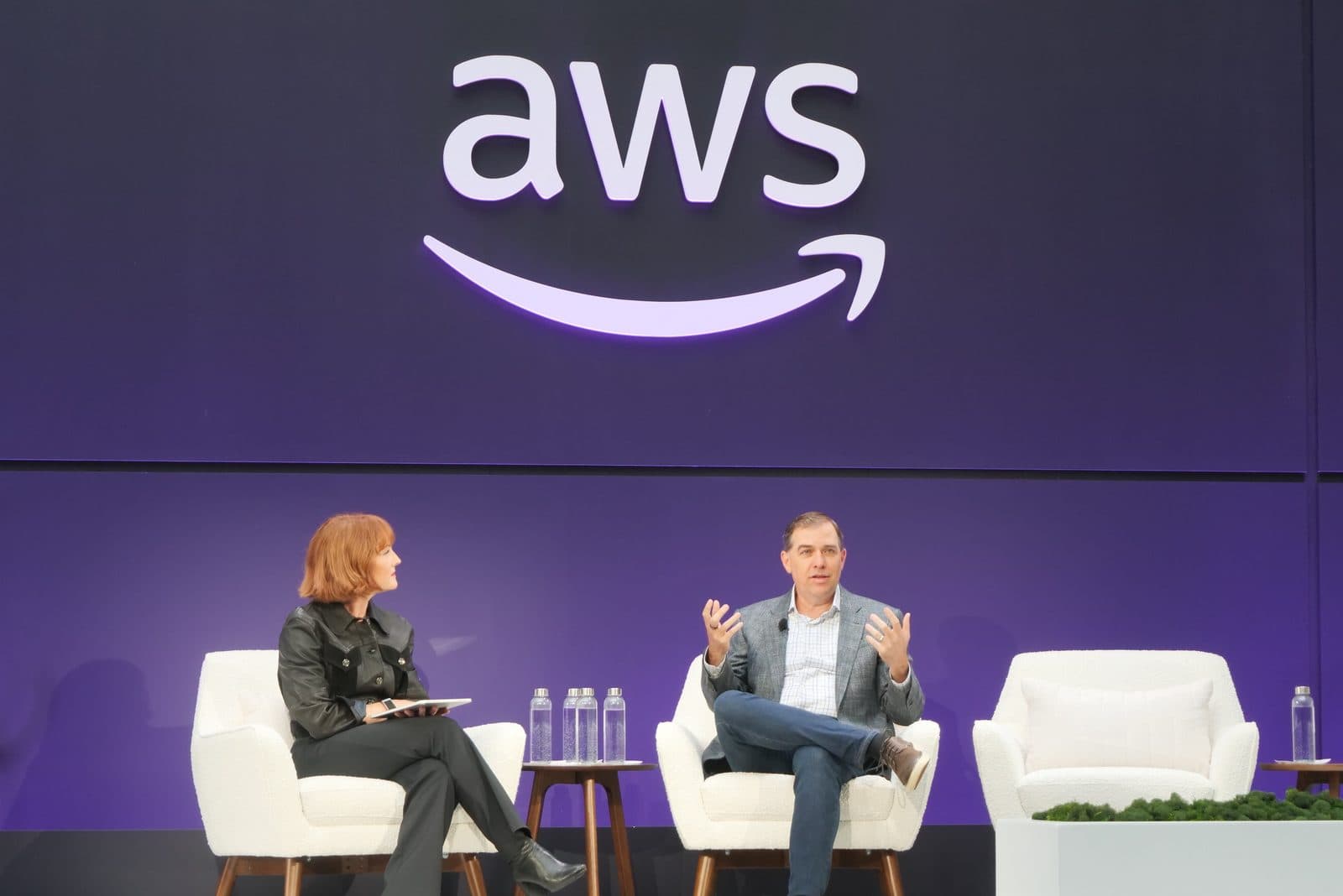 متحدثان جالسان على منصة أمام شعار AWS مضاء كبير في فعالية لـ Amazon Web Services.