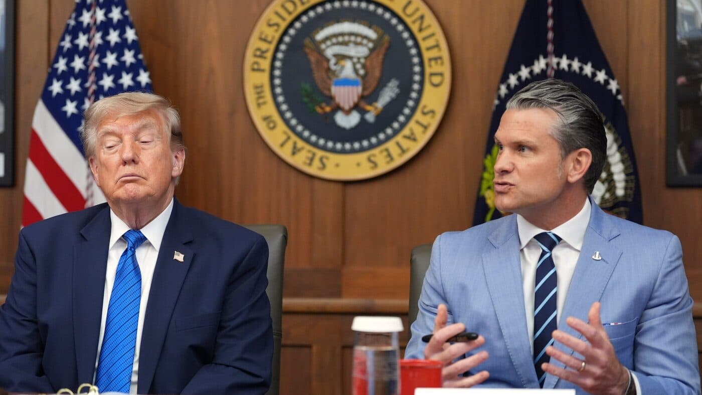 President Trump seated beside Defense Secretary Pete Hegseth in front of the presidential seal and US flags during a White House meeting