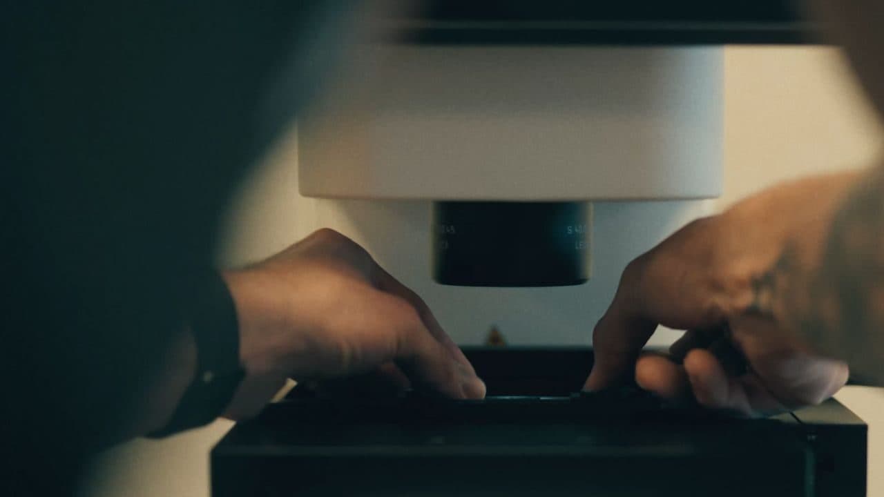 Close-up das mãos de uma pesquisadora operando um microscópio sobre uma bancada de laboratório em baixa luz