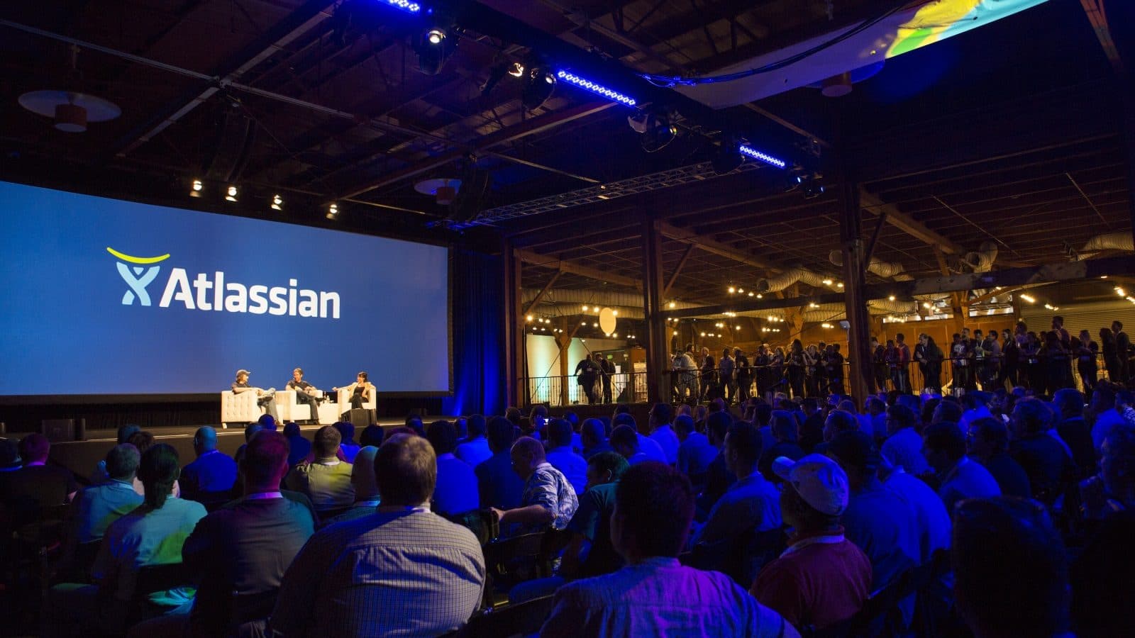 Un pubblico seduto in una conferenza, illuminato da una luce blu, di fronte a uno schermo del palco con il logo Atlassian