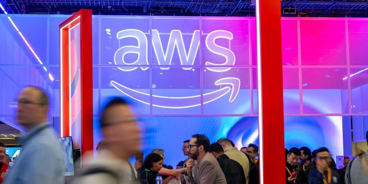 Conference attendees walking past a large illuminated AWS logo on a booth wall.