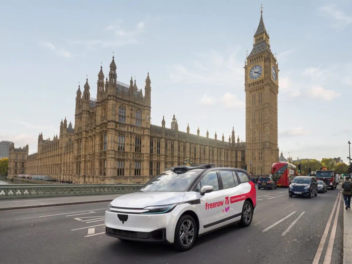 A white Freenow-liveried test car on the road beside the Houses of Parliament and the Elizabeth Tower in London, with black cabs and a red bus behind it