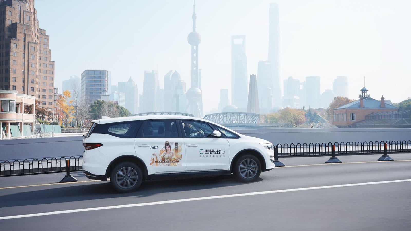 A white CaoCao robotaxi parked on a riverside road with the Shanghai skyline behind it