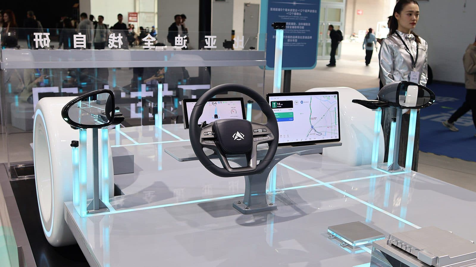 Autonomous-driving exhibit at a Chinese trade show, showing detached steering wheels and instrument displays mounted on a lit platform.