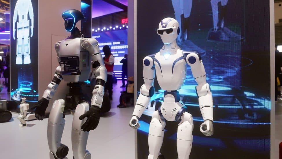 Two humanoid robots on display at a Chinese technology exhibition, one dark-shelled and one white, in front of an illuminated blue stand.