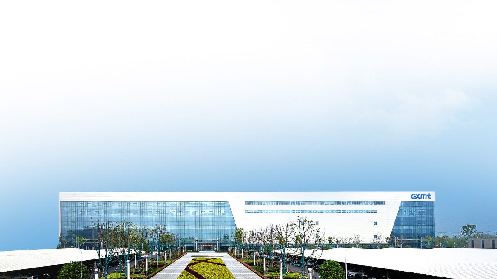 A wide view of a low semiconductor manufacturing campus at the end of a landscaped approach road under a pale sky.
