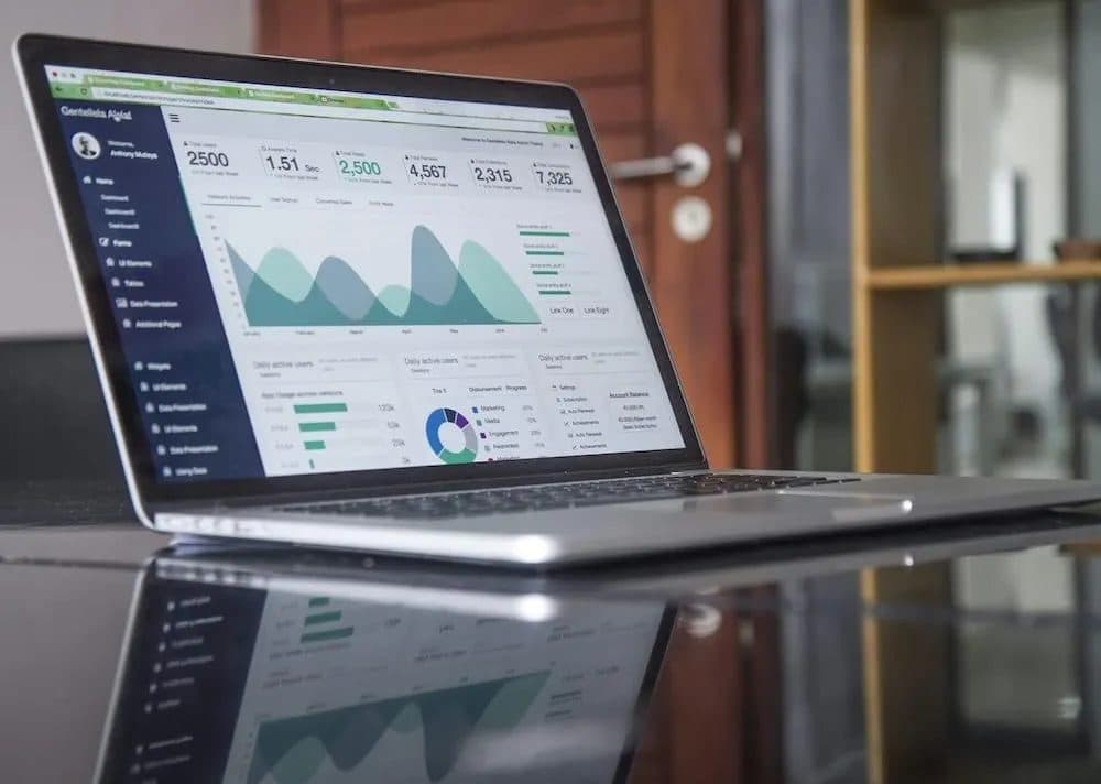 A stock photograph of an open laptop on a glass table displaying an analytics dashboard of charts, counters and activity graphs.