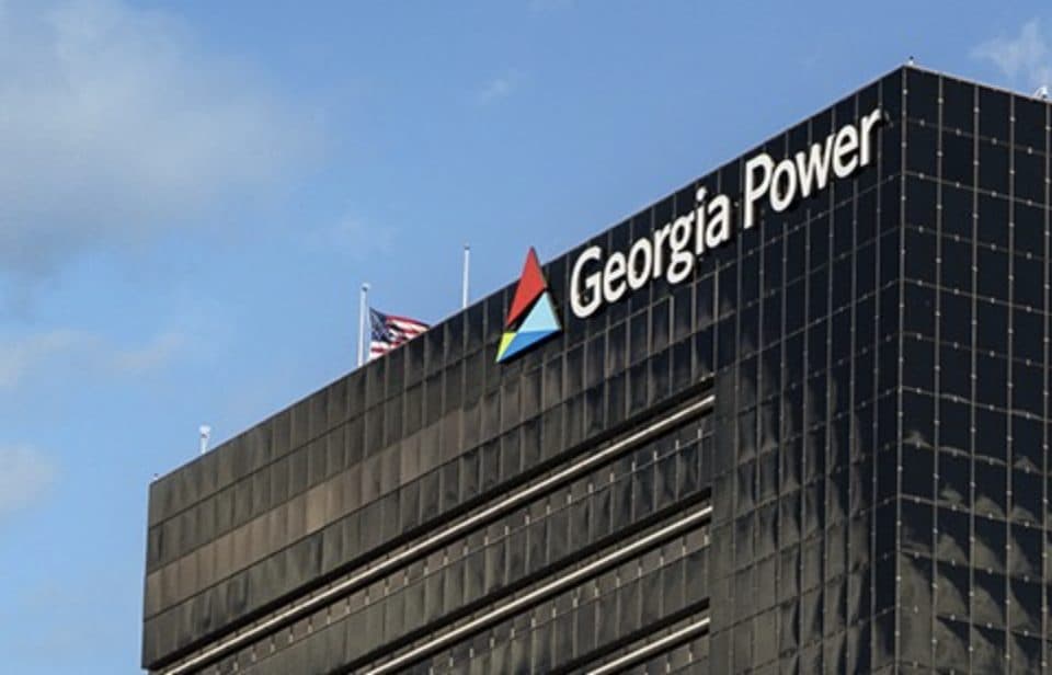 Os andares superiores de uma torre de escritórios de vidro escuro com o nome Georgia Power e o logotipo triangular, com uma bandeira dos EUA tremulando no telhado sob céu azul.