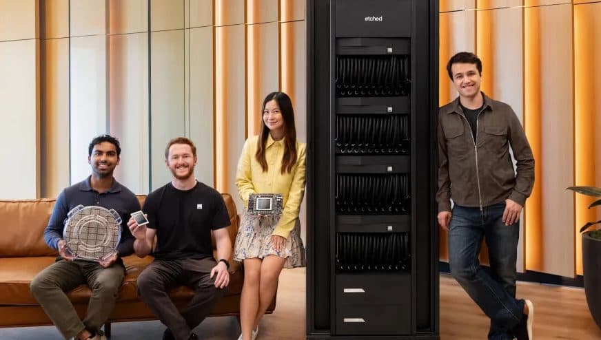 Quattro dipendenti di Etched accanto al rack di inferenza dell’azienda, con un wafer, un chip e una scheda