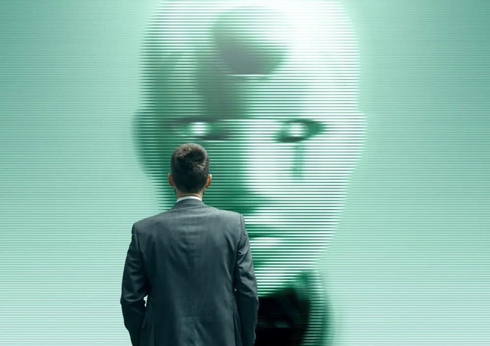 Um homem de terno visto de costas, diante de uma grande imagem borrada de um rosto humano renderizado em linhas de varredura verdes em uma tela.