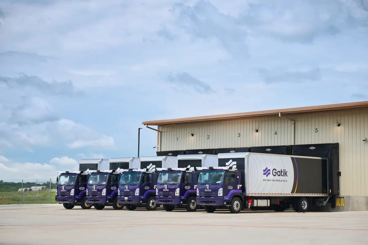 Uma fileira de caminhões-baú com a marca Gatik estacionados nas docas de carga de um armazém de distribuição