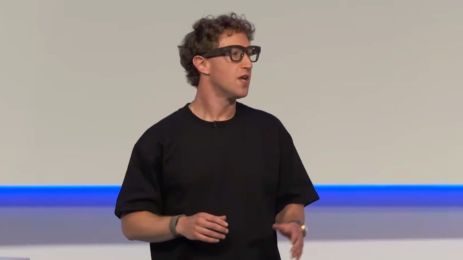 Un hombre con camiseta negra y gafas inteligentes, hablando en un escenario muy iluminado.