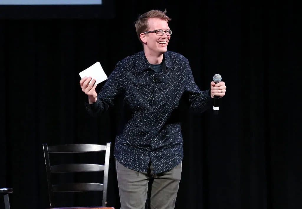 Hank Green on a darkened stage, holding a microphone in one hand and a sheet of paper in the other.
