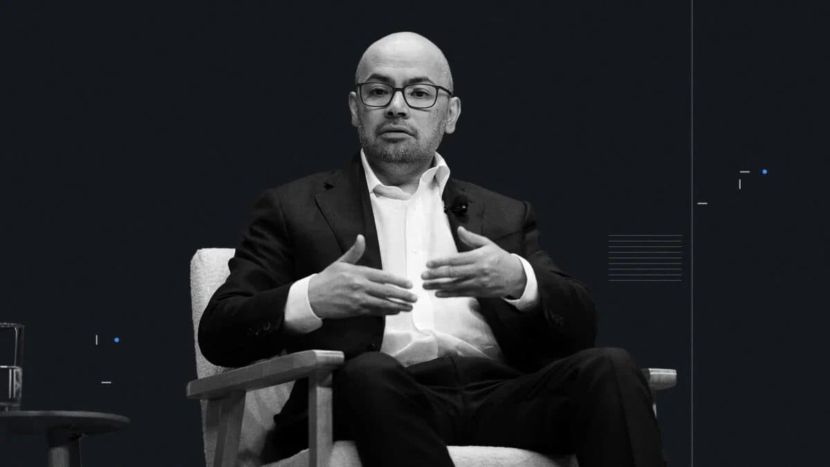 Demis Hassabis speaking from an armchair on a conference stage, in black and white