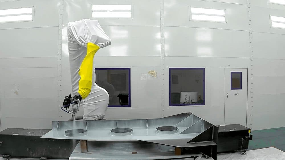 A worker in white protective coveralls leaning over a large steel panel inside a shipyard fabrication bay