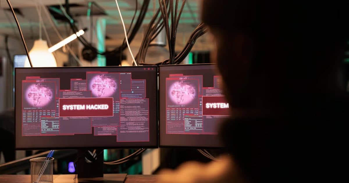 Силуэт человека в затемнённой комнате перед двумя мониторами с красными предупреждениями «system hacked».