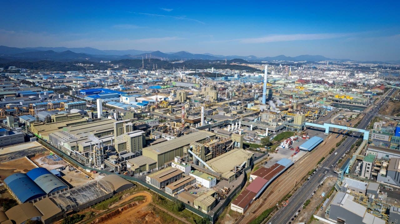 Uma vista aérea de um grande complexo industrial de fundição com prédios de processo de telhados azuis e tanques de armazenamento, ao fundo de colinas e água aberta.