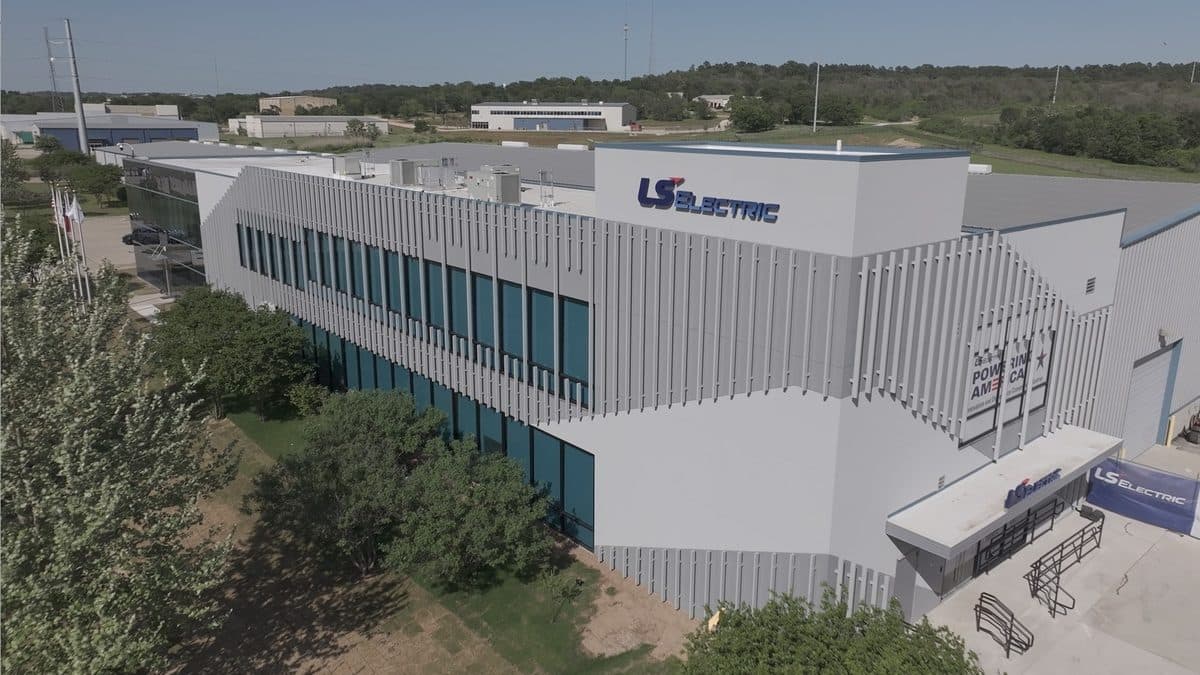 An aerial view of an LS Electric facility, a long low white building with the company's red logo on its side, surrounded by trees.