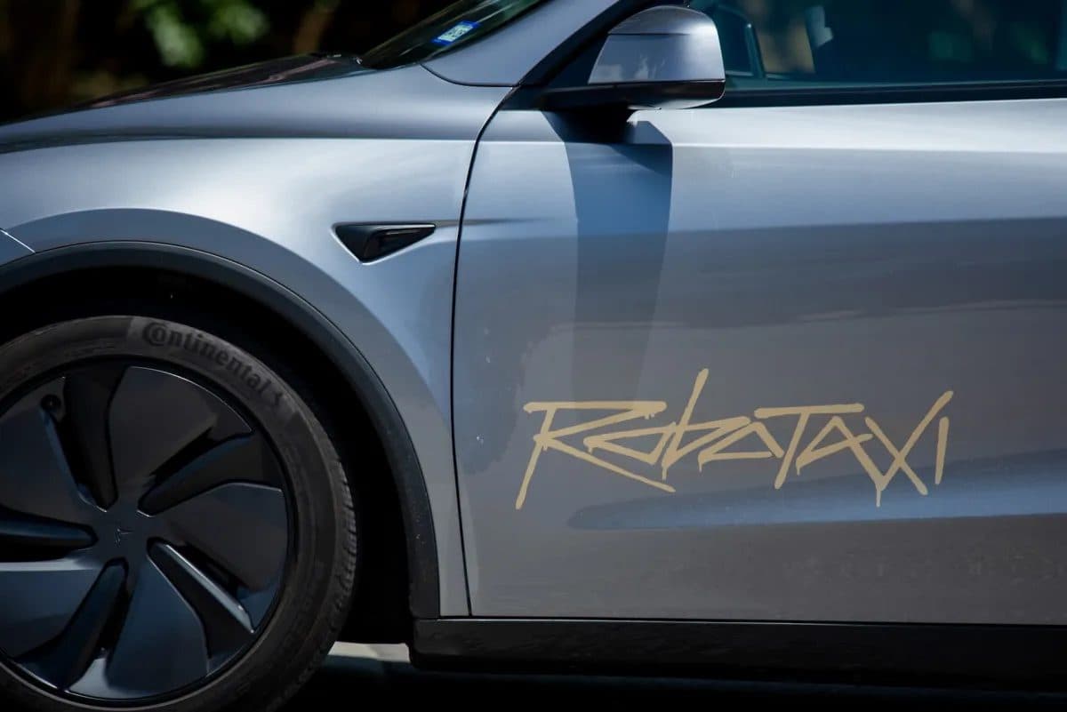 Side view of a silver Tesla vehicle with the word Robotaxi lettered along its rear door.