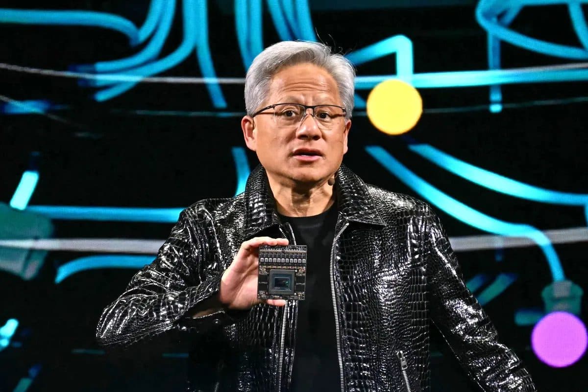 O CEO da Nvidia, Jensen Huang, em sua jaqueta preta de couro, segurando um chip para a plateia diante de um fundo de linhas curvas azuis.