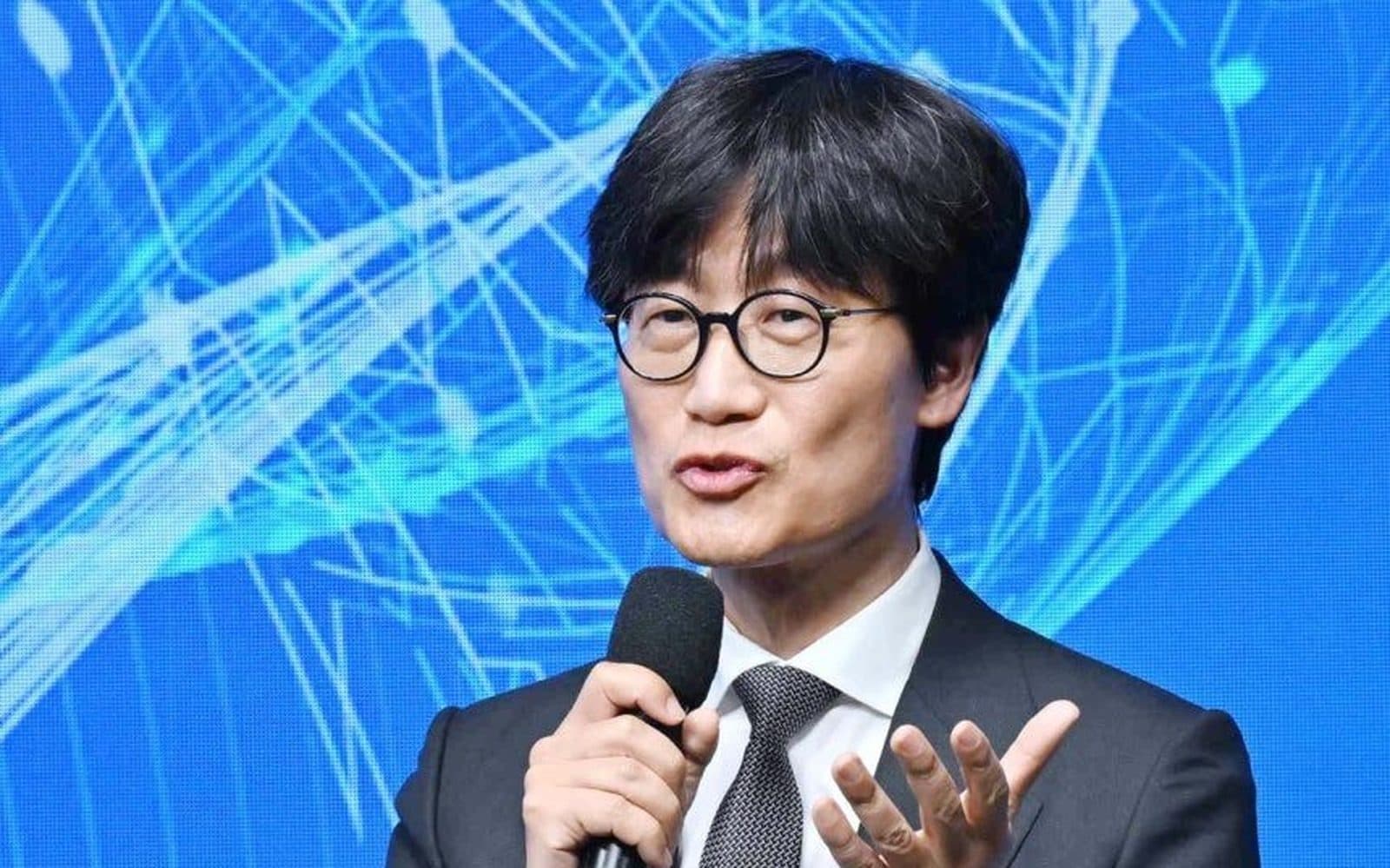 Un relatore con gli occhiali tiene un microfono a una conferenza, davanti a uno sfondo blu con grafica di rete
