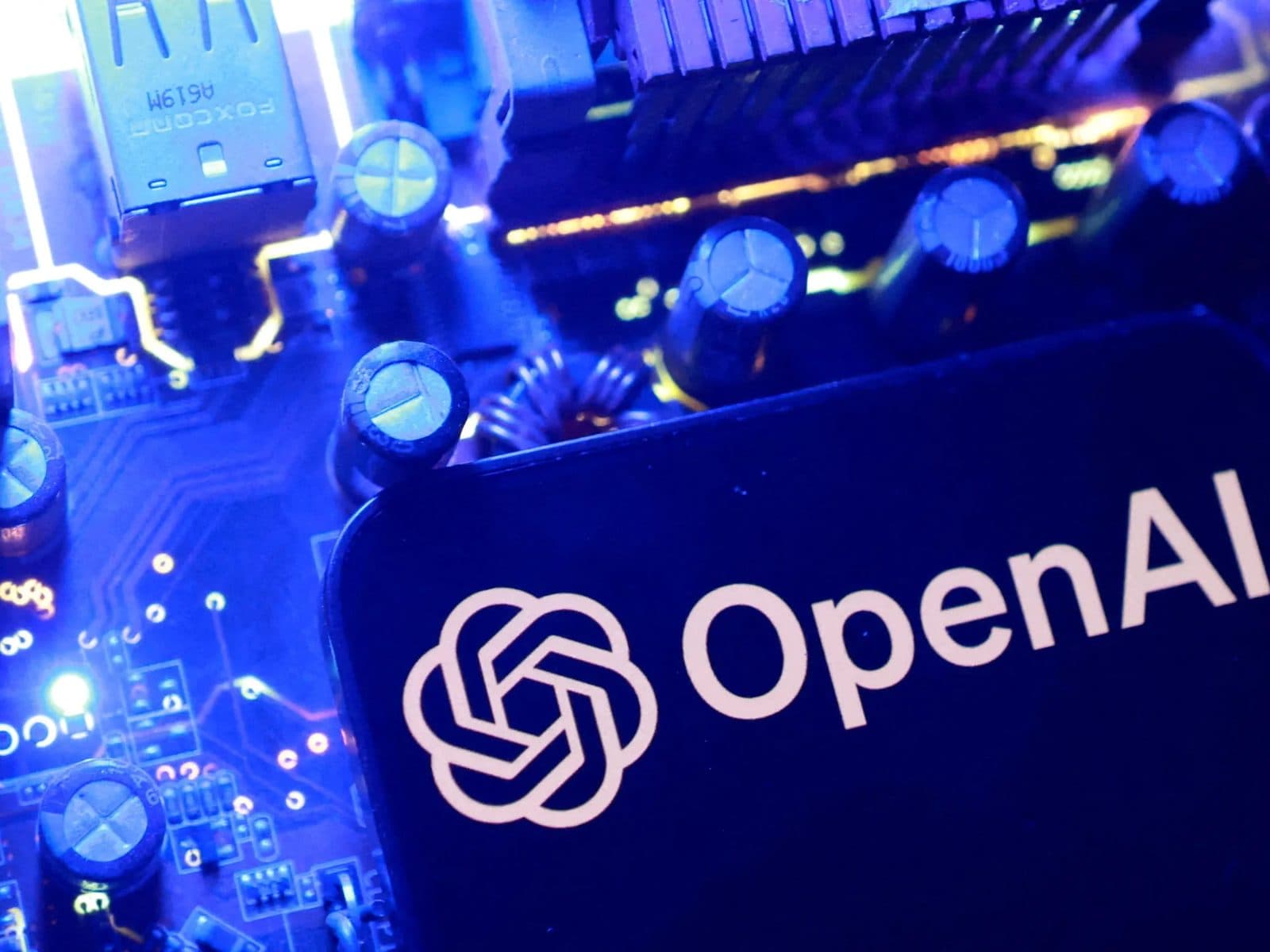 Le mot-symbole et le logo en spirale d’OpenAI sur une carte appuyée contre des circuits informatiques bleus illuminés