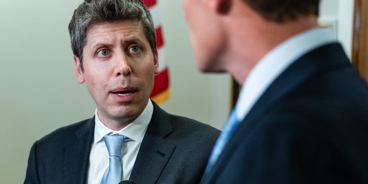 OpenAI chief executive Sam Altman, in a dark suit and pale blue tie, speaking to a man whose back is to the camera, with a United States flag behind him.