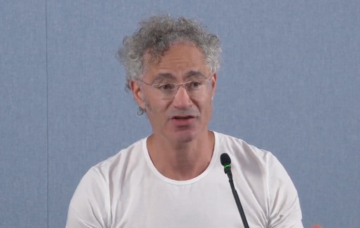 Palantir chief executive Alex Karp speaking into a microphone in a white T-shirt against a plain blue backdrop.