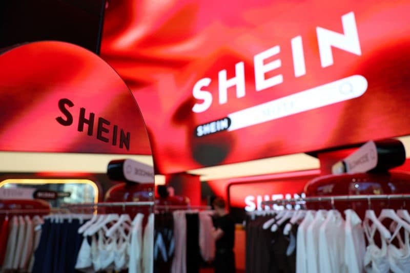Des sacs rouges arborant la marque SHEIN suspendus au-dessus d’une rangée de vêtements dans un magasin