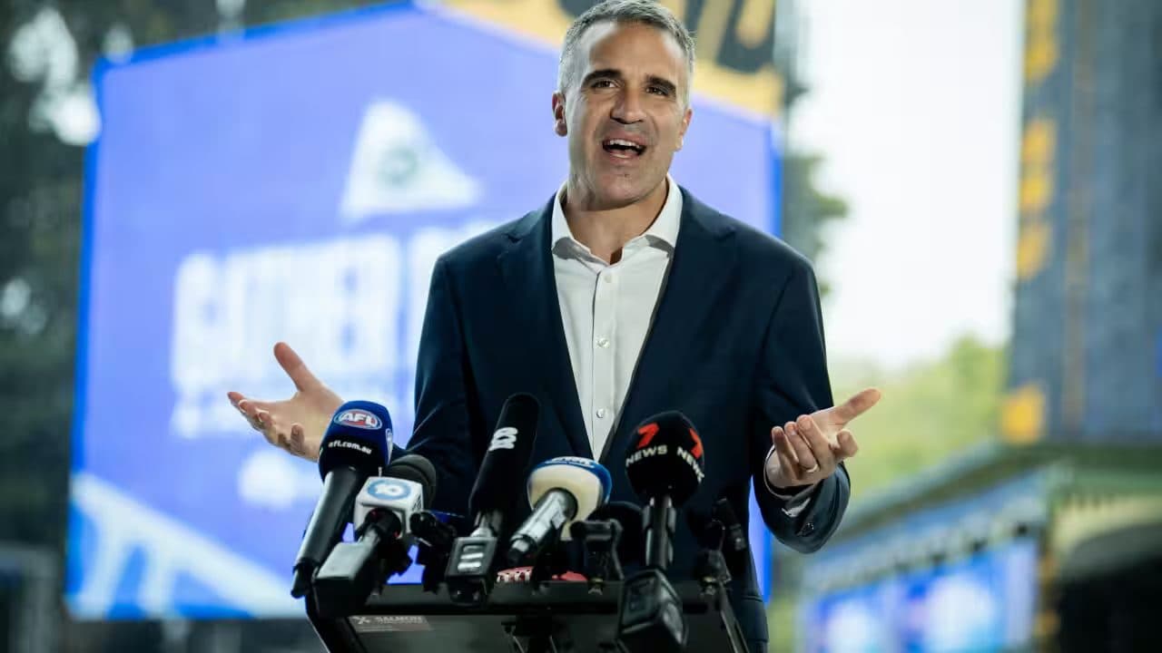 Il premier del South Australia Peter Malinauskas gesticola con entrambe le mani mentre parla davanti a una fila di microfoni televisivi, con uno schermo blu sullo sfondo.