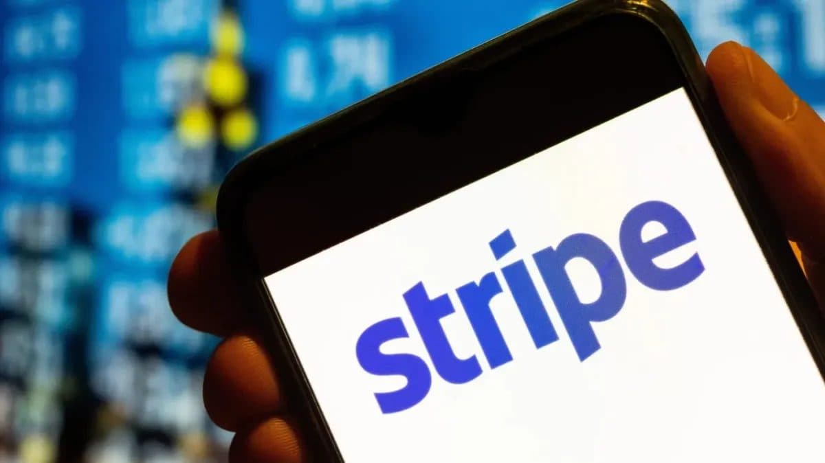 A smartphone showing the Stripe wordmark, held in front of an out-of-focus screen of financial market data.