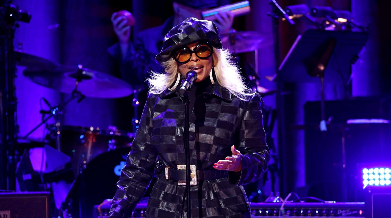 Mary J. Blige che si esibisce al microfono con giacca e cappellino in pelle scozzese e occhiali da sole rotondi tinta, sotto le luci del palco