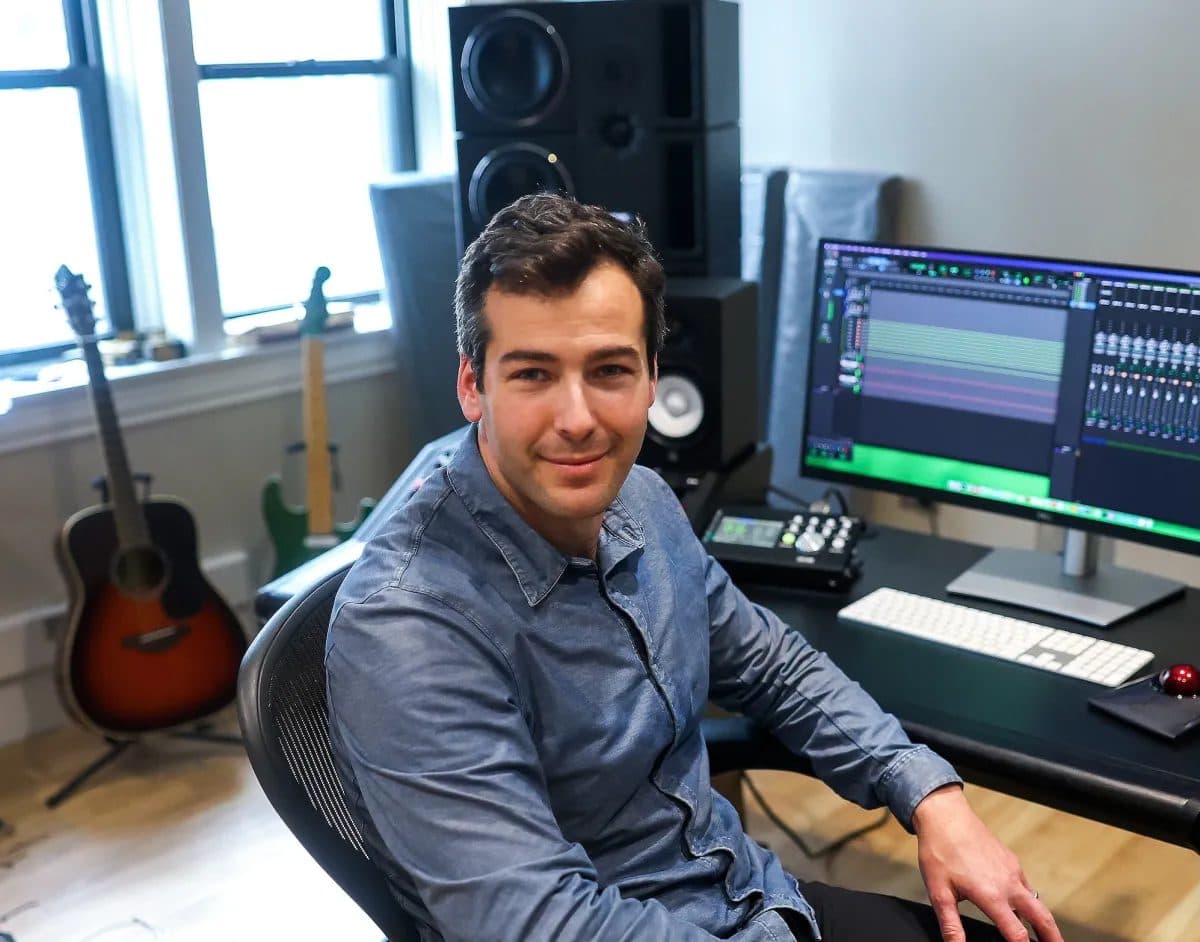 A man seated at a home studio desk with studio monitors, a keyboard and an acoustic guitar beside him