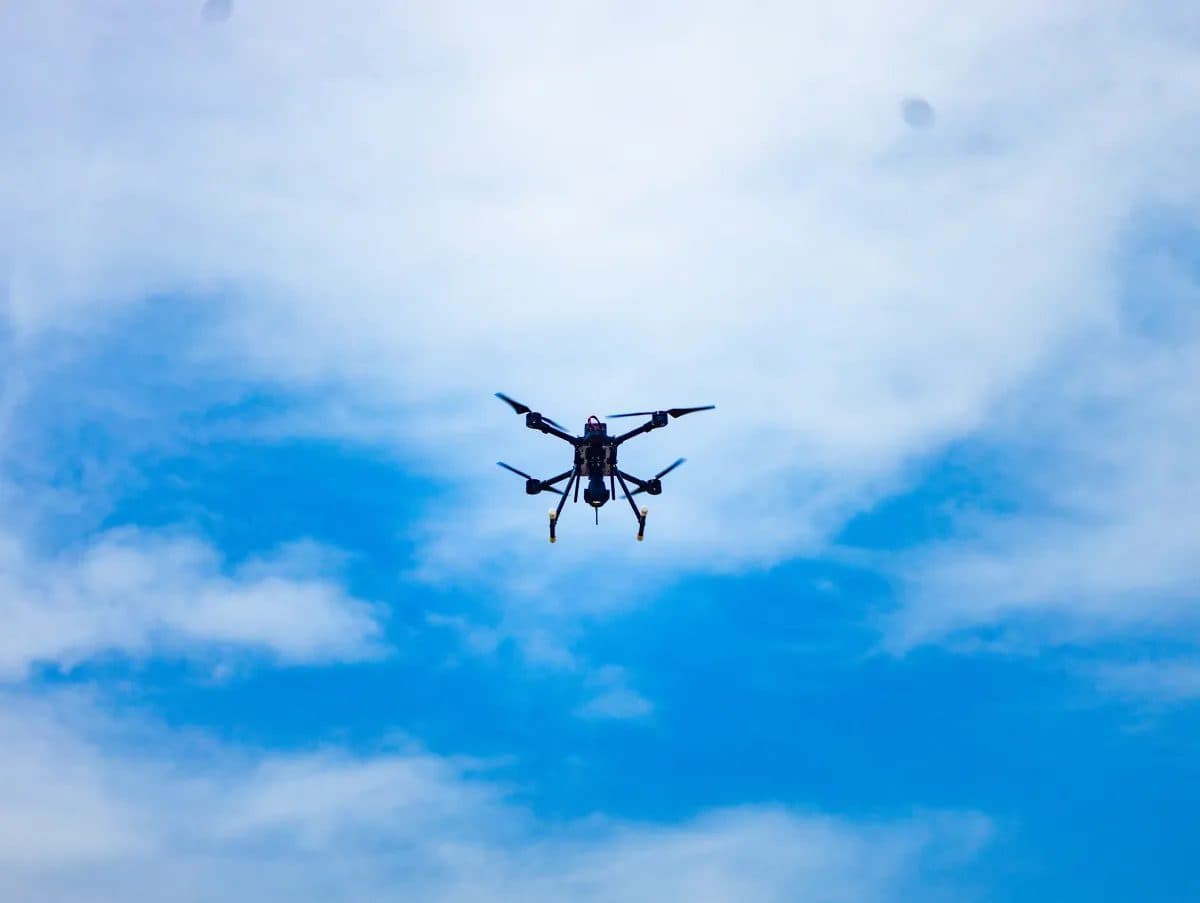 A small quadcopter drone silhouetted against a blue sky with light cloud