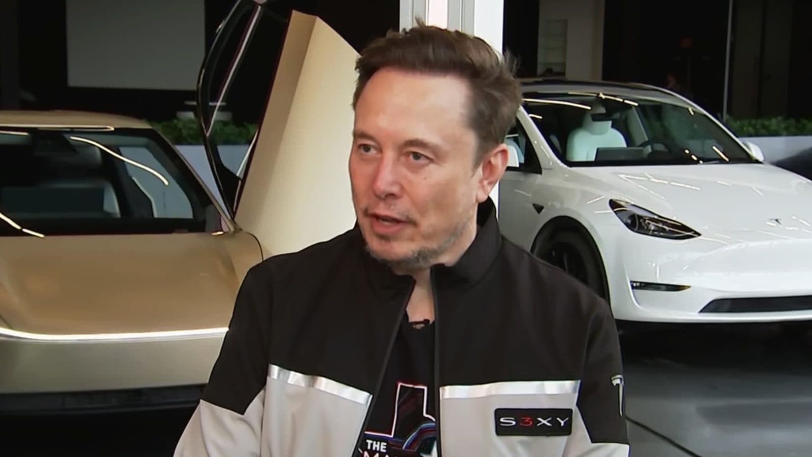Elon Musk يتحدث في صالة عرض مع سيارات Tesla خلفه