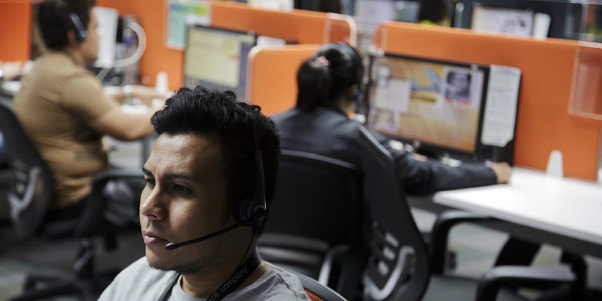 Call-centre workers wearing headsets at computer workstations in an open-plan office, one man in the foreground mid-call.