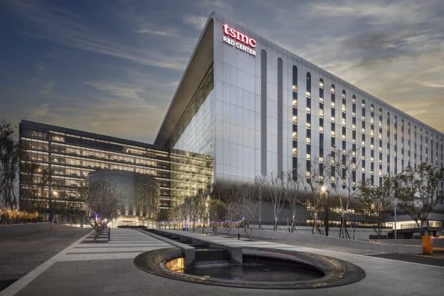 Esterno di un edificio TSMC al tramonto, con facciata in vetro e cemento che curva sopra un piazzale paesaggistico e il logo dell'azienda vicino alla linea del tetto.