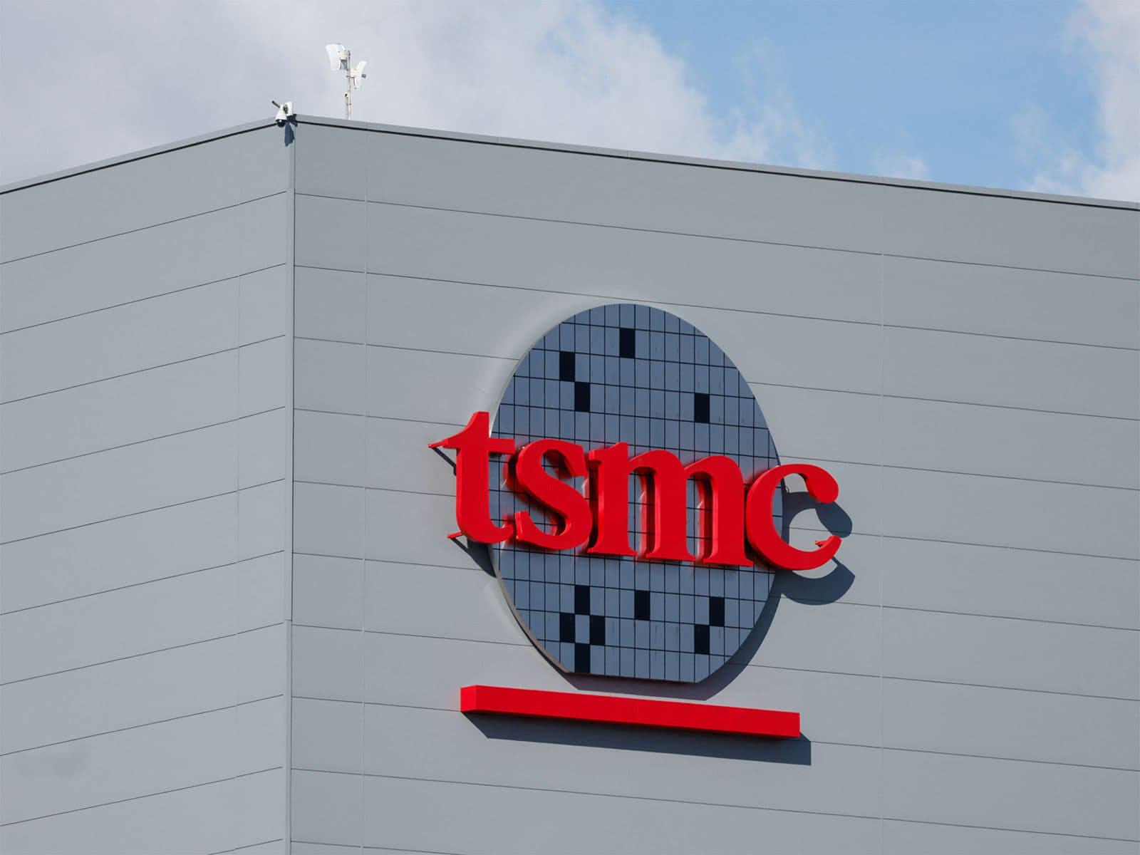 The TSMC wordmark and wafer logo mounted on the grey exterior wall of a company building against a blue sky.