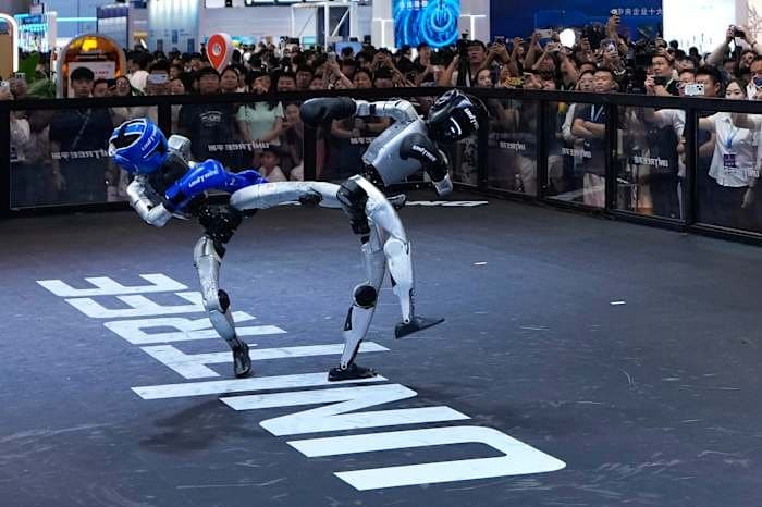 Two Unitree humanoid robots in blue and white boxing headgear sparring in a Unitree-branded ring before a crowd filming on phones.