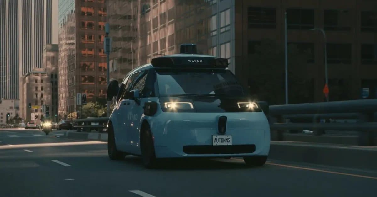 A driverless Waymo vehicle with roof-mounted sensors on a city street.