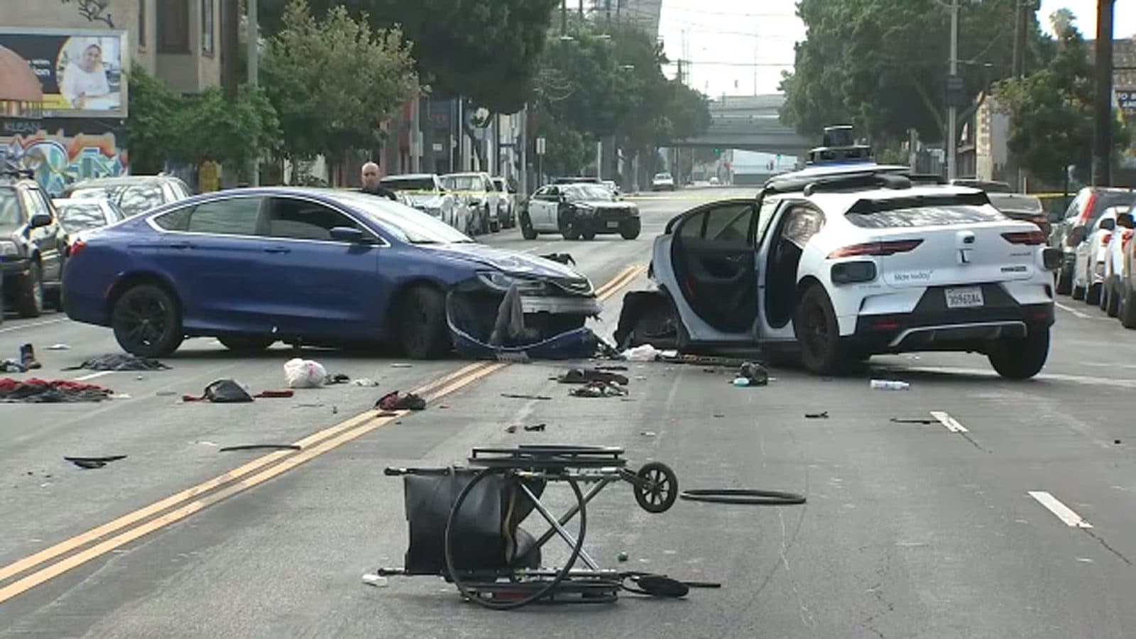 La scène d’accident dans une rue de Los Angeles : une berline sombre endommagée, une Waymo blanche avec une porte ouverte, et des débris, dont un chariot renversé, sur la chaussée