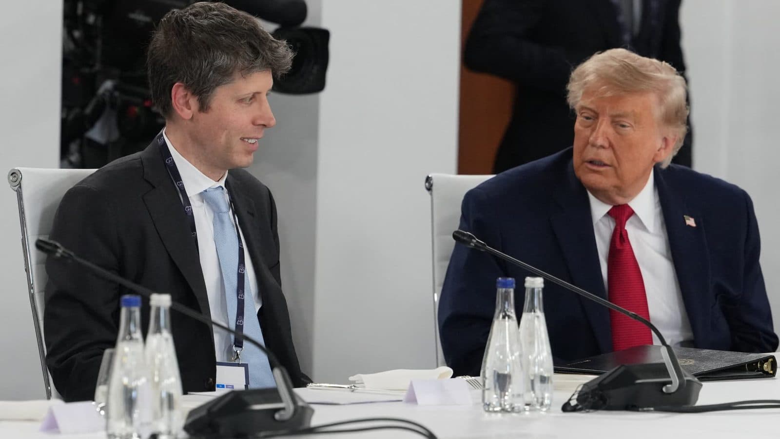 OpenAI chief executive Sam Altman seated beside President Donald Trump at a roundtable, with microphones and water bottles on the table.
