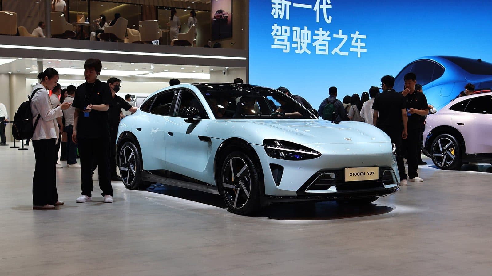 Un crossover elettrico Xiaomi YU7 azzurro chiaro su un espositore in showroom, con visitatori e personale tutt'intorno.