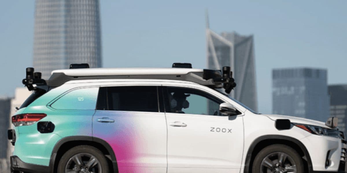 A white Zoox-branded SUV with roof-mounted sensor housings parked on a city street, of the retrofitted type used for mapping runs.
